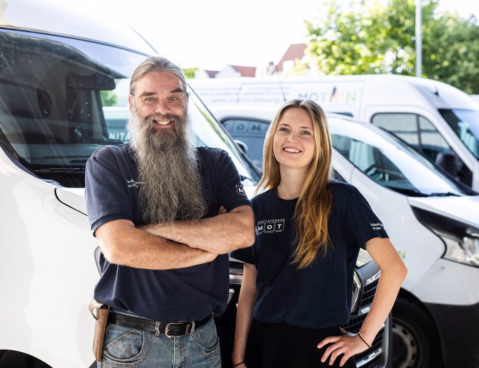 Service-Team von MOT vor Firmenfahrzeugen Zwei MOT-Mitarbeitende stehen nebeneinander vor weißen Firmenfahrzeugen, beide in Arbeitskleidung und freundlich lächelnd